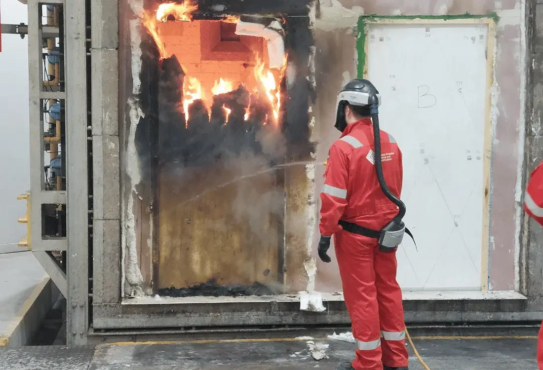 Fire door undergoing a controlled fire resistance test in a laboratory, with flames visible through the door while a technician in protective safety gear monitors and sprays water during the assessment.